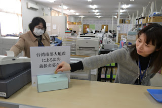 町役場の受け付けカウンターに設置された「台湾南部大地震による災害義援金募金」と書かれた水色の募金箱に、カウンターの中の女性が募金を入れ、もう一人の女性がその様子を見ている写真