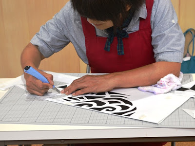 赤いエプロンを着た女性が細かい模様の黒と白の切り絵をカッターで切っている様子の写真