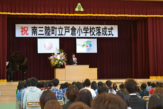 体育館の壇上に花と日の丸と町章の幕が飾られ、多くの児童が見つめる中で一人の男性が演台であいさつしている落成式の写真
