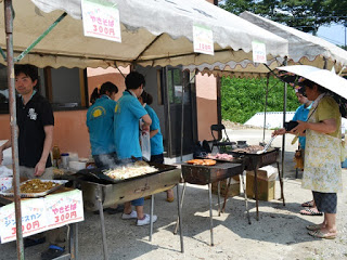 白いテントの下に3つの焼き台がおかれ、水色のシャツを着たスタッフが焼きそばやジンギスカンの準備をしており、その前には買い物をする女性の姿が見える写真