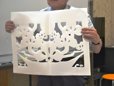 年配の男性が教室の中で大きな白い紙の松の切り絵を両手で持ち上げて見せている写真