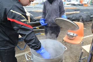 湯気をあげる寸胴鍋の後ろで一人が蓋を持って立ち、横に立ったもう一人が、ゴム手袋をつけた手を鍋に入れ、温度を測っている様子の写真
