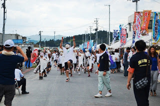 沿道の人々が見守っているなか、先頭のお面をつけた踊り手のグループが両手を上げて勢いよく通りを進んでいる様子の写真