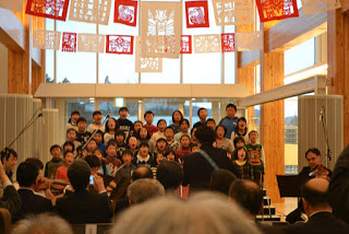 木の梁と大きな窓がある屋内で、天井からきりこの飾りが吊るされている中、階段に並んで立った小学生の合唱団が観客の前で歌っている様子が写っている写真