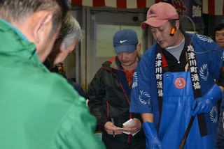 エプロンと手袋を着け法被には「志津川町漁協」と書かれている人物の周りに、複数の人物が集まっている競りの様子の写真