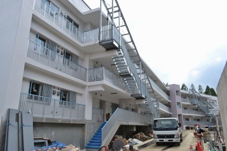 完成に近い三階建ての共同住宅に外階段が取り付けられ、建物の前には工事資材が並べられている写真