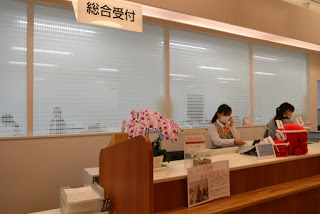 カウンターの左端にピンク色の蘭の鉢植えと募金箱が置かれ、後ろでは二人の女性職員が仕事をしている、南三陸病院の総合受付の写真