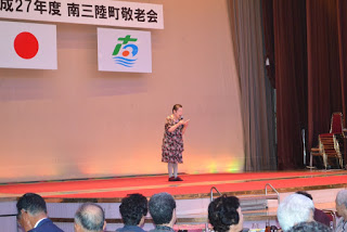 舞台の上で柄のワンピースを着た高齢の女性が一人でマイクを持って歌っており、客席の人々が見守っている写真