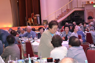 会場内で立ち上がった高齢の女性が、テーブルに座る他の人たちに笑顔で話しかけている様子の写真