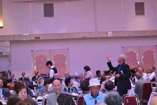 会場内でスーツ姿の高齢の男性が立って手を挙げており、まわりには着席した参加者や配膳中のスタッフが見える写真