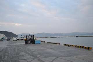 曇り空の下、遠くの水面越しに山々が望める背景に複数の車両や建物が見える海辺のコンクリートの岸壁にフォークリフトがあり、水色のコンテナを運んでいる、港の作業場の写真