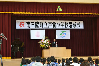 舞台に設置された演台でスーツの男性がマイクの前に立ち、壇上左右にピアノと花が飾られている小学校落成式の写真