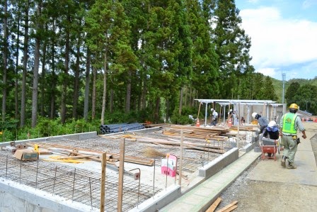 林のそばの平地で基礎工事が行われており、鉄筋や木材が置かれて複数の作業員が作業をしている写真