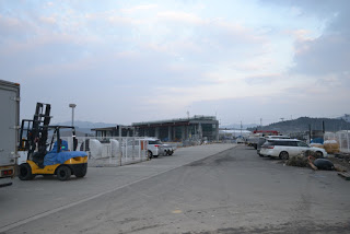 空はところどころ雲がかかっていて、遠くには山々が見える左側にフォークリフトがあり、周囲に複数の車が駐車され、背景には建物が見える漁港の写真