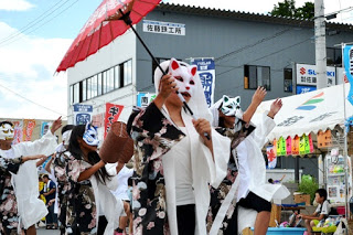 狐のお面をつけ、白い羽織を着た踊り手たちが赤い和傘を差しながら、妖艶な雰囲気で通りを練り歩いている様子の写真