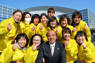 お揃いの黄色のジャンバーを着たスタッフの方12名がスーツ姿の男性を中心に記念撮影をしている写真