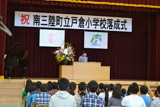 児童たちが椅子に座って見守る前で、青い服を着た女性が演台に立ち、マイクで話をしている南三陸町立戸倉小学校落成式の写真