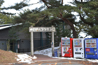 松の木の奥にある建物の前に木で作られた「神割観光プラザ入口」と書かれたゲートがあり、その右手前に3台の自動販売機が並んでいる写真