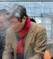 湯気越しに写した、薄茶色のジャケットに赤と黒の配色のマフラーを首に巻いた眼鏡をかけた男性の写真