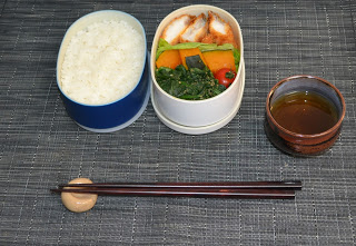 楕円形の2段のお弁当箱の一つに白ご飯、もう一つにカボチャや青野菜、揚げ物のおかずが盛り付けられ、お茶と箸が添えてある写真