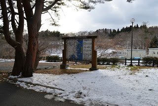 雪が残る地面に、地図などの案内が載った大きな看板が立っており、その左側に松の木が生えている写真