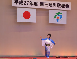 舞台の上で青と白の着物を着た女性が一人で日本舞踊を踊っている様子で、背後には町章と日の丸が掲げられている写真