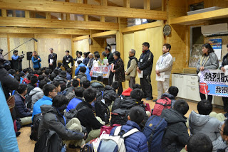 前方に歓迎の横断幕を持った人など、たくさんの人が一列に並んで立ち、その前で荷物を背負った大勢の学生たちが床に座っている会場の様子の写真