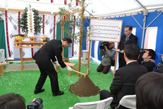 坂川所長がテントの中に設置された神棚の前で、緑色の敷物の上に盛られた土に鍬を入れている写真