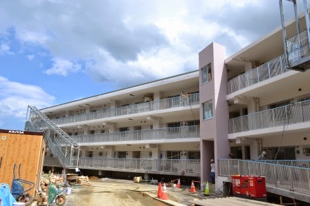 三階建ての共同住宅の端にあるピンク色のエレベーター塔のある建物が、整備途中の駐車場とともに写されている写真