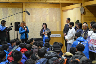 室内でたくさんの子どもたちが集まって座り、前方に立つ数人の大人がマイクを使って話している様子の写真