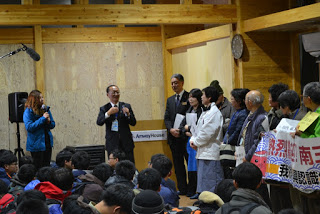 座った学生たちの前に立った代表の男性が、歓迎する住民らに挨拶をしている様子の写真