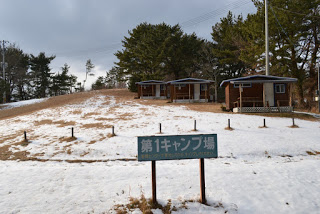 「第1キャンプ場」の看板の奥に、木々に囲まれた広々とした広場があり、右側に同じつくりの家が3棟並んで立っている写真