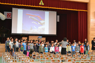 体育館の舞台前に整列した子どもたちが、指揮をとる先生の前で合唱しており、後方のスクリーンにはこいのぼりの写真が映し出されている合唱発表の写真