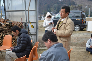 開け放たれたハウスの入り口横にまきが積まれており、中にならんだオレンジ色の椅子に2人の男性が座り、入り口付近に男性と女性が立っている様子の写真