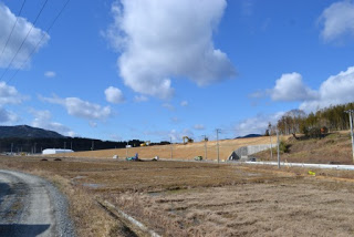なだらかな斜面の手前に車が走る道路が通っている様子を手前の空き地越しに撮影した写真