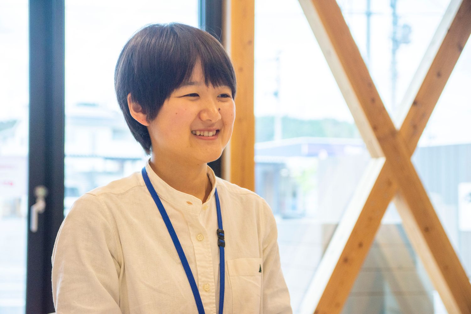 図書館の窓際で、白いシャツを着た女性が青いストラップの名札を首から下げて笑顔で話している様子をアップで撮影した写真