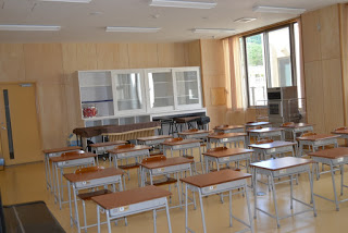 大きな窓から自然光が差し込む、木の内装が明るい教室に、誰もいない状態で整然と机と椅子が並べられている教室内の写真