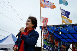 風に揺れる旗を背景に、マイクを持って話している亜KIRAさんの写真