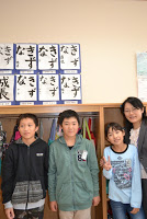 教室の棚の前に立つ三人の児童と一人の女性教師が、笑顔で並んでおり、棚の上には習字で「きずな」と書かれた作品が掲示されている記念撮影の写真