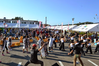 白いテントが並んだ屋外の広場で、観客が見守るなか、大勢の人がそろいの衣装で元気に踊っている写真