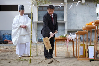 白い装束を着た神主さんが見守る中、スーツ姿の男性が鍬入れの儀で砂を掘っている様子の写真