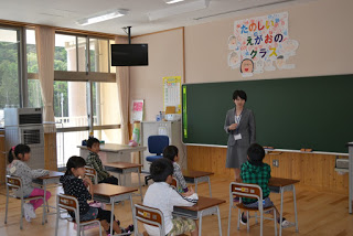 黒板の前に立つ女性教師が、机に座った児童たちに話しかけており、「たのしい えがおの クラス」と書かれた掲示が壁に貼られている授業風景の写真