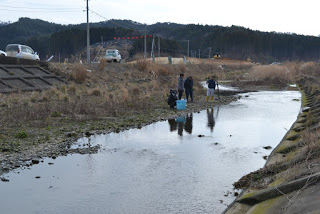 奥に森が広がる川のほとりに水色のバケツが置かれ、4人の人物が集まっている写真