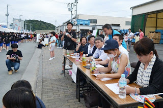 道路脇に設けられた審査員席で、大人たちが真剣な表情で踊りを見ながら審査用紙に記入している様子の写真