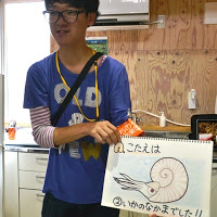 青いシャツを着た男性が「こたえは2.いかのなかまでした！！」とイラストとともに書かれたスケッチブックを持って立っている写真