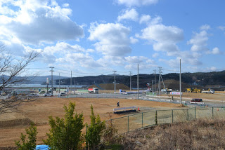 電柱が立ち並ぶ道路の左右に土の空き地があり、工事車両などが出入りしている様子を高台から撮影した写真