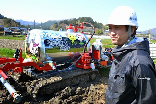 イラストが描かれた銀色のタンクを搭載した赤い液肥専用散布車の前に、安全帽をかぶった森谷さんが立っている写真