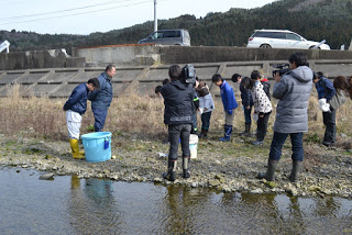 川岸に水色の大きなバケツが置かれ、その隣に立った男性2人と、向かい合って立った子どもたちがお辞儀をしている様子を、ビデオカメラを持った男性2人が撮影している様子の写真