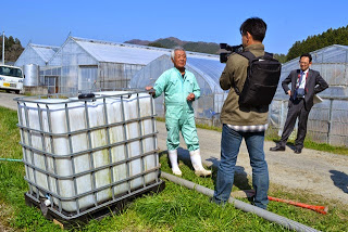 格子枠の台に置かれた大型タンクの前で、緑色の作業服を着た白髪の男性にカメラを向け、向かい合って立つ男性から取材を受けている様子の写真