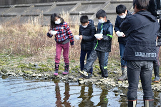 白いお椀型の容器を手に持ち、ゆっくりと歩いている4人の子どもたちと、その様子を川の中から撮影している男性の写真